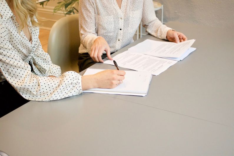 Negotiation - woman signing on white printer paper beside woman about to touch the documents