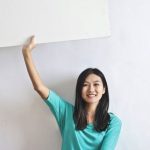 Tech Real Estate - Cheerful Asian woman sitting cross legged on floor against white wall in empty apartment and showing white blank banner