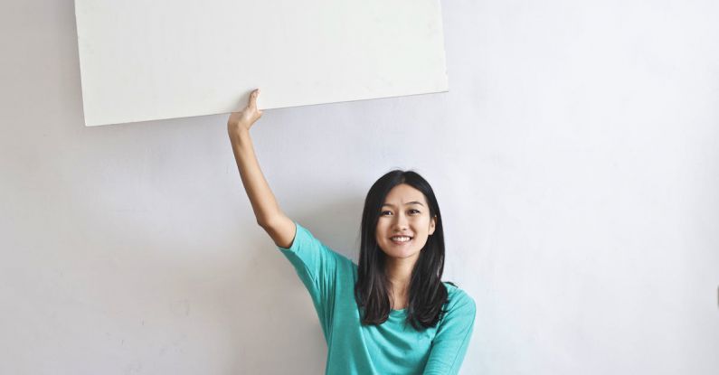 Tech Real Estate - Cheerful Asian woman sitting cross legged on floor against white wall in empty apartment and showing white blank banner