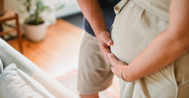 Future Home - From above of crop faceless couple in casual outfit standing in light living room and touching pregnant belly gently