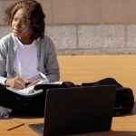 Online Platform - Contemplative African American schoolkid with copybook and netbook sitting with crossed legs on wooden platform while looking away in sunlight